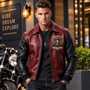 Op de afbeelding: Een bordeauxrode en zwarte leren varsity-jas met een kraag, ritssluiting aan de voorzijde en geribbelde boorden. Op de linkerborst zit een geborduurde patch met een adelaarsymbool en de tekst BROOKLYN ATHLETIC CLUB EST. 2027. Twee zakken met rits.