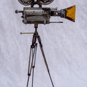 Könnte beinhalten: Eine Skulptur einer alten Filmkamera aus recycelten Metallteilen, darunter Kugellager für die Spulen und ein gelb lackierter Metalltrichter als Objektiv. Die Kamera ist auf einem Stativ aus dünnen Metallstangen und Schrauben montiert.