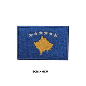 May include: A rectangular embroidered patch featuring the flag of Kosovo with a blue background, a gold map of Kosovo in the center, and six white stars arranged in an arc above the map. The patch measures 9 cm by 6 cm as indicated by text.