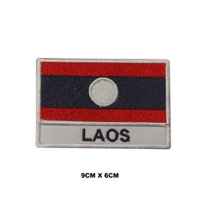 May include: A rectangular embroidered patch featuring the flag of Laos with red and dark blue horizontal stripes and a central white circle. Below the flag, a light gray bar displays the text LAOS in black. The patch measures 9cm by 6cm.