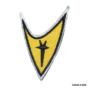 May include: An embroidered patch in the shape of a stylised arrowhead, featuring a yellow centre with a black vertical emblem and a white and black border. The patch measures 9.8 cm by 6 cm, as indicated by the text in the corner.