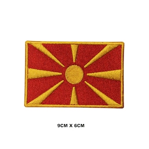 May include: A rectangular embroidered patch featuring the North Macedonian flag design with a red background and a central yellow sun with eight radiating sunbeams. The patch measures 9 cm by 6 cm as indicated by the text below the item.