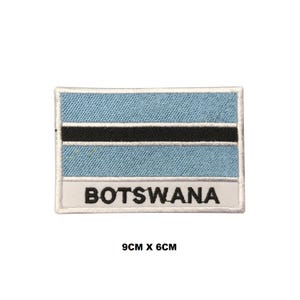 May include: A rectangular embroidered patch featuring the flag of Botswana with light blue, white, and black horizontal stripes. The word BOTSWANA is stitched in black capital letters on a white background at the bottom. It measures 9cm x 6cm.
