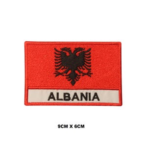 May include: A rectangular embroidered patch featuring a red background with a black double-headed eagle emblem centred above a white horizontal stripe containing the black text ALBANIA. The patch measures 9 cm by 6 cm.