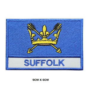 May include: A rectangular embroidered patch featuring a blue background with a yellow crown and two crossed arrows. Below the emblem, a white horizontal band displays the text SUFFOLK in blue letters. The patch measures 9cm by 6cm.