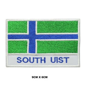 May include: A rectangular embroidered patch featuring a green field with a blue cross outlined in white. Below the cross, a white horizontal band contains the blue text SOUTH UIST. The patch measures 9 cm by 6 cm as indicated by the text.
