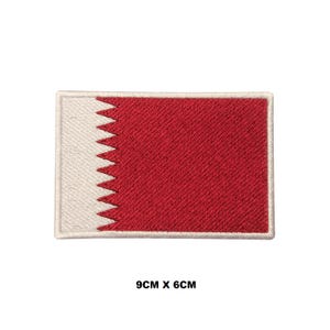 May include: A rectangular embroidered patch featuring the flag of Qatar with a white vertical band on the left side and a larger red section on the right, separated by a serrated edge with nine points. The patch measures 9 cm by 6 cm.