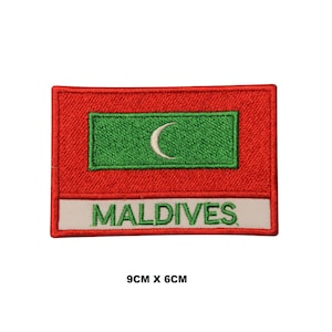 May include: A rectangular embroidered patch featuring the flag of the Maldives with a red border, a green center rectangle, and a white crescent moon. Below the flag, a white horizontal bar displays the text MALDIVES in green, measuring 9cm x 6cm.
