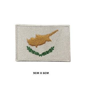 May include: A rectangular embroidered patch featuring the flag of Cyprus with a white background, a gold map of the island, and two green olive branches underneath. The patch measures 9 cm by 6 cm as indicated by the text below the item.