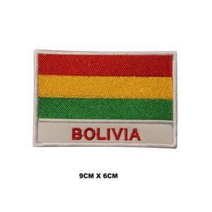 May include: A rectangular embroidered patch featuring the horizontal tricolor flag of Bolivia with red, yellow, and green stripes. Below the flag, a white horizontal band displays the word BOLIVIA in red capital letters. Dimensions are 9cm x 6cm.