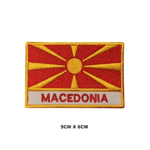 May include: A rectangular embroidered patch featuring the flag of North Macedonia with a yellow sun and eight rays on a red background. A white horizontal bar at the bottom displays the red text MACEDONIA. The patch measures 9cm by 6cm.