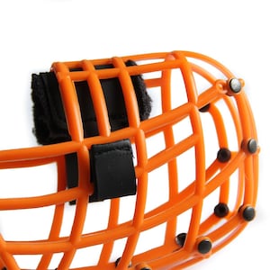 May include: Close-up of an orange protective cage with a black strap and fasteners. The cage has a grid-like structure and is designed to protect the face. The fasteners are small, black, and round.