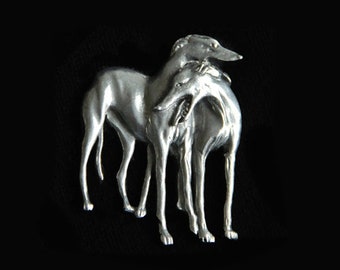 May include: A silver-toned metal sculpture of two greyhounds, one atop the other, set against a black background. The dogs are depicted in a stylized, elegant pose, showcasing their slender forms and refined features. The sculpture is likely a decorative item.