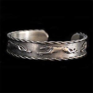 Könnte beinhalten: Ein silbernes Armband mit einem geflochtenen Design und drei laufenden Greyhound-Silhouetten um das Band.