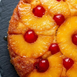 Könnte beinhalten: Ein runder Ananas-Umkehrkuchen mit goldbraun karamellisierten Ananasscheiben auf der Oberseite. Jede Ananasscheibe ist in der Mitte mit einer leuchtend roten Maraschino-Kirsche verziert, die einen Kontrast zur glasierten Oberfläche bildet.