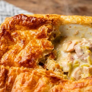 Puede incluir: Un pastel salado con una corteza de hojaldre dorada y crujiente. El interior revela una salsa blanca cremosa con trozos visibles de pollo cocido y puerros en rodajas, con vapor saliendo del plato caliente recién horneado.