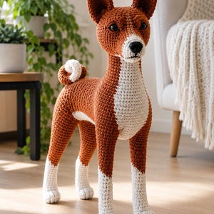 May include: A crochet dog figurine featuring a rust-brown body with white markings on the chest, muzzle, and lower legs. The stuffed animal has upright pointed ears, dark plastic eyes, a black nose, and a tightly curled tail positioned on its back.