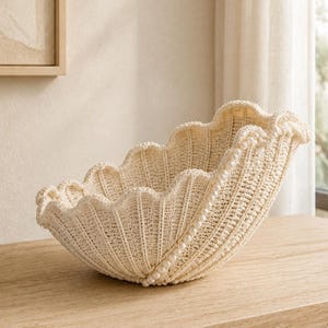 May include: A cream-coloured crocheted bowl with a scalloped edge and a ribbed texture. The bowl is embellished with a single strand of small white faux pearls that follows the curve of the rim, adding a decorative detail to the textile construction.