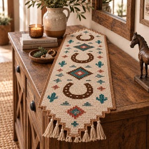 May include: A rectangular table runner featuring a cream-coloured woven fabric with a cross-stitch pattern of brown horseshoes, teal cacti, and geometric diamond shapes in red and teal. The runner has a brown border and cream-coloured tassels.