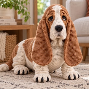 Puede incluir: Una figura de perro Basset Hound tejida a ganchillo con un patrón tricolor de hilo marrón, blanco y crema. El peluche tiene orejas largas y caídas, una nariz cosida en negro y ojos oscuros, sentado en una pose realista con detalles negros.