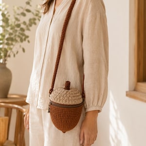 May include: A crochet crossbody bag shaped like an acorn, featuring a dark brown base and a textured light tan cap. The bag includes a matching dark brown crochet shoulder strap and a dark metal zipper closure at the top with a small stem detail.