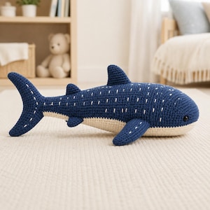 May include: A handmade crochet whale shark plush toy featuring a deep blue upper body with white stitched dot patterns and a solid cream-coloured underbelly. The stuffed animal has a prominent dorsal fin, side fins, and a wide tail fin.