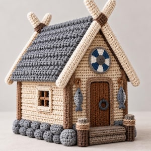 May include: A handcrafted crochet house featuring a dark grey textured roof, beige walls, and a stone-look base. The front includes a brown door with a ring handle, two hanging grey fish, a blue and white lifebuoy emblem, and wooden-look posts.