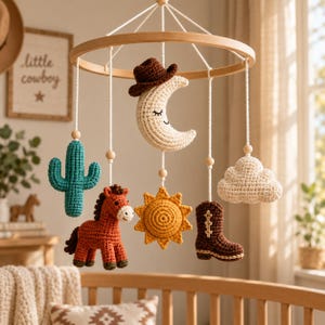 May include: A nursery mobile featuring crocheted figures suspended from a circular wooden frame. The figures include a cream crescent moon wearing a brown cowboy hat, a teal cactus, a brown horse, a yellow sun, a brown boot, and a cream cloud.