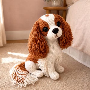 Könnte beinhalten: Ein handgefertigtes Häkel-Stofftier in Form eines Cavalier King Charles Spaniel Hundes. Das Plüschtier hat ein weiß-braunes Farbmuster, große schwarze Kunststoffaugen, eine schwarze Nase sowie lange, strukturierte, lockige braune Garnohren und einen Schwanz.