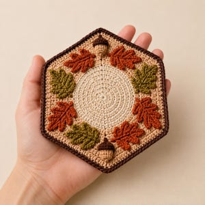 May include: A hexagonal crochet coaster featuring a central beige circle surrounded by a ring of alternating dark green and burnt orange leaves, accented with two small brown acorns at the top and bottom, all bordered by a dark brown yarn edge.