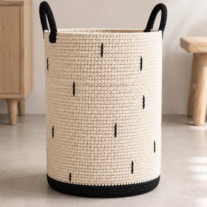 May include: A cylindrical woven basket featuring a cream-colored body with scattered vertical black stitch accents. The basket includes two sturdy black handles at the top and a solid black base, constructed with a consistent crochet-style texture.