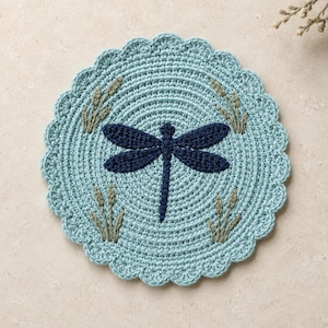 May include: A round, light blue crocheted coaster featuring a dark navy blue dragonfly centered in the middle. The coaster has a scalloped edge and is decorated with small, embroidered light green wheat-like stalks in each of the four corners.