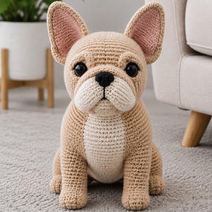 May include: A crochet French bulldog stuffed animal featuring tan yarn with a cream-colored chest and muzzle, large upright ears with pink inner detailing, and black plastic eyes. The dog is sitting in an upright position with a black nose.