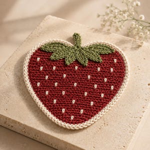 Può includere: Un sottobicchiere all'uncinetto a forma di cuore che ricorda una fragola. Il corpo principale è rosso scuro con piccoli dettagli di semi ricamati in bianco. Presenta un gambo e foglie verdi sulla parte superiore, bordati da un profilo crema.