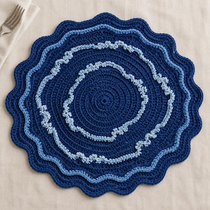 Op de afbeelding: Een ronde, gehaakte placemat met concentrische cirkels van diep marineblauw garen. Het ontwerp bevat twee golvende, lichtblauwe, getextureerde ringen die een gelaagd effect creëren, met een geschulpte randafwerking in bijpassend marineblauw.