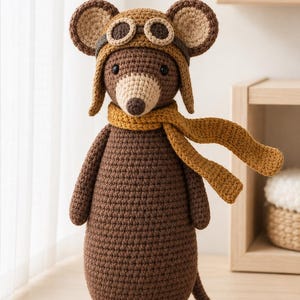 Puede incluir: Un ratón de ganchillo marrón que lleva un gorro de aviador marrón oscuro con gafas de color beige y una bufanda de color amarillo mostaza. El ratón tiene un hocico beige claro, ojos negros pequeños y una cola larga. Hecho con puntos visibles.