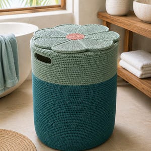 May include: A cylindrical storage basket made of woven rope with a two-tone design featuring a dark teal base and a light sage green top. The lid is shaped like a flower with sage green petals and a textured coral pink center, including two side handles.