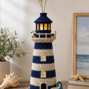 Op de afbeelding: Een gehaakt vuurtorenbeeldje met horizontale marineblauwe en crèmekleurige strepen, een grijze voet en een blauw golfaccent. De structuur bevat een verlichte lantaarn, kleine ramen en twee kleine vogelfiguren op de reling van de galerij.