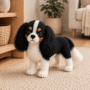 May include: A crocheted stuffed animal in the likeness of a Cavalier King Charles Spaniel dog. The toy features black, white, and tan yarn, with long black ears, a white blaze on the face, dark brown eyes, and a black nose, standing on four legs.