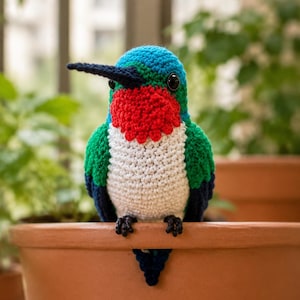 May include: A crocheted hummingbird figurine featuring a bright blue head, a vibrant red throat patch, a white chest, and green and dark blue wings. The bird has a long, thin, dark beak and small black eyes, perched with dark feet visible.