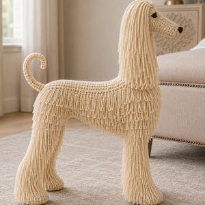 May include: A cream-colored crocheted Afghan Hound figurine featuring long, flowing yarn strands that mimic the breed's signature coat. The dog has a slender neck, a curled tail, and dark, stitched eyes and nose, crafted entirely from yarn.
