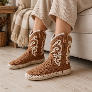 May include: A pair of mid-calf boots crafted from brown crochet yarn, featuring a cream-coloured sole and intricate cream-coloured stitched embroidery patterns along the sides. The boots have a pull tab at the top and a rounded toe design.