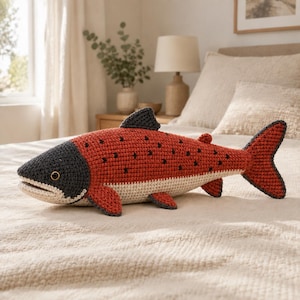 May include: A crocheted stuffed fish toy featuring a dark gray head, a rust-orange body with small black spots, and a cream-colored underbelly. The fish has a prominent dorsal fin, pectoral fins, and a large tail fin, all crafted with yarn.
