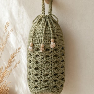 May include: A sage green crochet bag with a cylindrical shape and a drawstring closure. The bag features a repeating shell stitch pattern, a top hanging loop, and three drawstrings finished with light wooden beads and small white seashells.