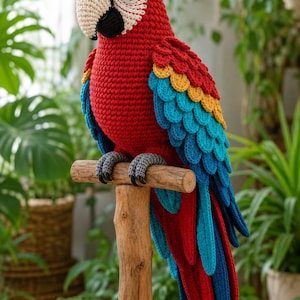 May include: A crocheted macaw parrot toy with a bright red body, yellow and blue wing feathers, and a cream-colored beak. The bird is perched on a wooden stand featuring a cylindrical post and a circular base, with dark gray crocheted feet.