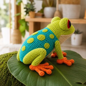 May include: A crocheted frog toy featuring a bright green head and limbs, a teal back with light green circular spots, and vibrant orange feet. The frog has a cream-colored underbelly and large eyes with black pupils and red accents on the sides.
