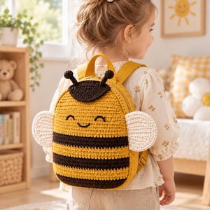 May include: A yellow and brown crocheted backpack shaped like a bee, featuring a smiling face, two black antennae, and two white crocheted wings on the sides. The backpack has a zip closure and adjustable yellow straps for carrying on the back.