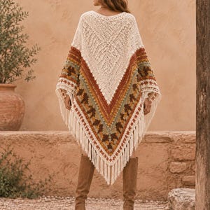 May include: A cream-coloured crochet poncho featuring a V-shaped design with horizontal bands of burnt orange, olive green, and brown geometric patterns. The garment has a textured knit construction and is finished with long cream fringe along the hem.