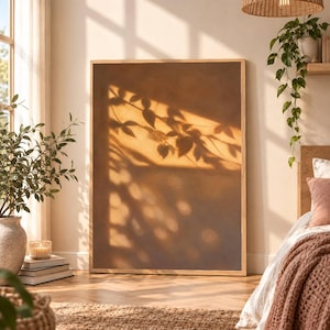 Könnte beinhalten: Ein großes, rechteckiges Wandbild in einem hellen Holzrahmen mit einem warmen, einfarbigen braunen Hintergrund und einem weichen, goldfarbenen Schattenwurf von Baumzweigen und Blättern auf der Oberfläche, der einen natürlichen Effekt erzeugt.