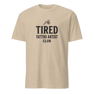 May include: A beige short-sleeve t-shirt featuring a black graphic of a tattoo machine above the text TIRED TATTOO ARTIST CLUB printed in a bold, sans-serif font. The garment has a crew neckline and a standard fit, showcasing a clean design.